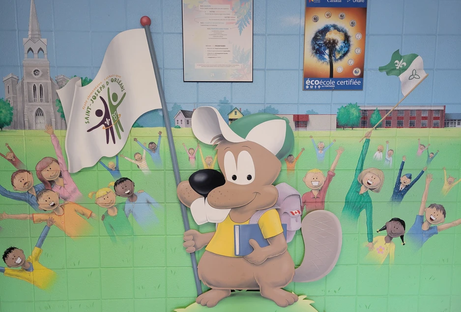 Murale colorée à l'entrée de l'école montrant la mascotte de SJO tenant un drapeau et entourée d'enfants joyeux.